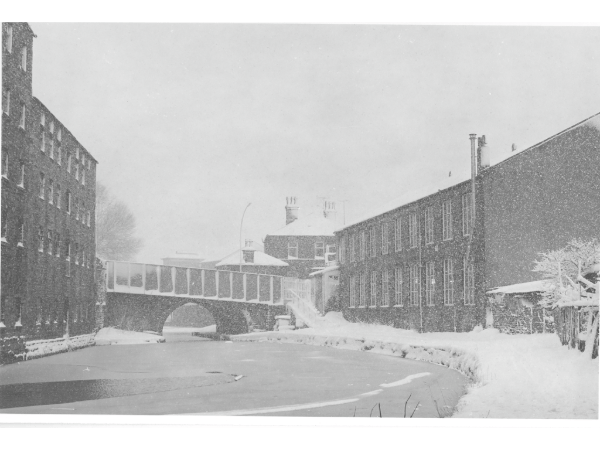 A view of Manchester Road Canal Bridge taken during the winter when the water was frozen and snow covered the tow path and the roofs of the houses and the mills.