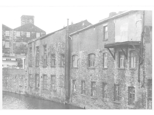 Photograph shows the rear of the first public library in Burnley, the Marshall Branch Library, which was opened in 1914. It was situated in what was known as Ruskin Hall on Trafalgar Street. The library can be seen in the middle of the image. The right hand side building is the Trinity Conservative Club. Both buildings have now been demolished.