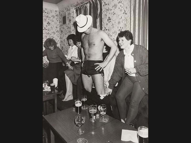 Bringing down the house! A group of cheerful friends share uncontrollable giggles during a lively November 1986 hen party at an unknown location, as a fedora-wearing entertainer strikes a dramatic pose right in the middle of the lounge. Surrounded by pints of Banks's Mild and a room full of roaring laughter, the fun-loving partygoers make sure the bride-to-be gets a send-off to remember!