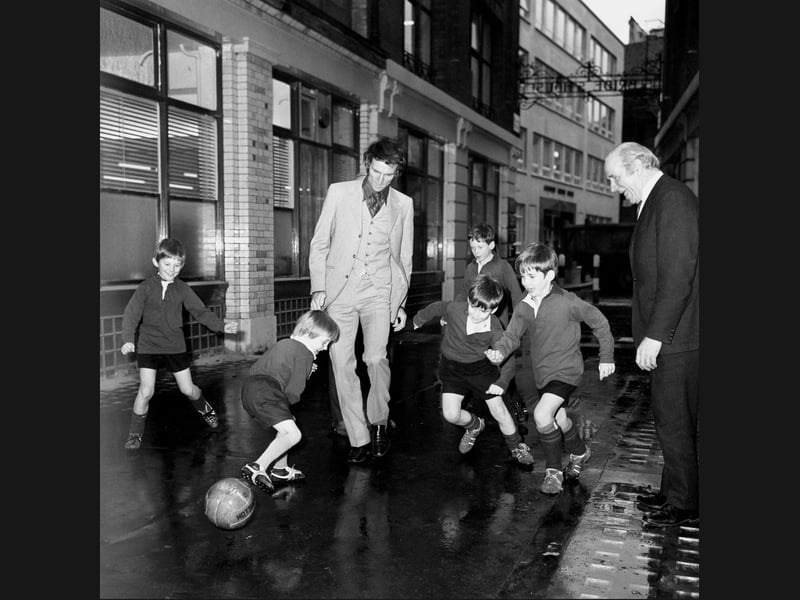 The ultimate kickabout! Wolverhampton Wanderers legend and PFA Chairman Derek Dougan joins Manchester United’s Sir Matt Busby for a game with eager young schoolboys outside the Press Club on November 19, 1970. Dressed in a sharp three-piece suit, "The Doog" displays his signature charm and footwork as he navigates the improvised pitch alongside the enthusiastic kids. The delightful street session captures the beloved Wolves star sharing his passion for the beautiful game off the pitch.