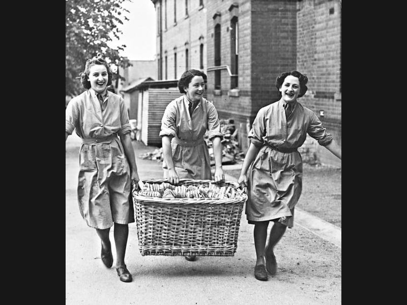 Bringing plenty of cheer to the mess hall! On May 22, 1940, three radiant Auxiliary Territorial Service orderlies share beaming grins as they haul a giant wicker basket overflowing with sliced bread. Clad in their working overalls, the cheerful trio prove that even routine wartime kitchen duties were delivered with wonderful spirits and absolute camaraderie.