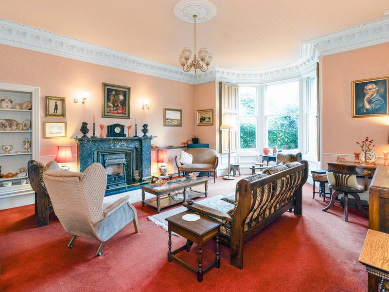 The principal living room is illuminated by a wide shuttered bay window framing leafy frontgarden views. An imposing marble fireplace with a living flame fire anchors the sitting area, complemented by cornicing, a ceiling rose, and an open Edinburgh press.