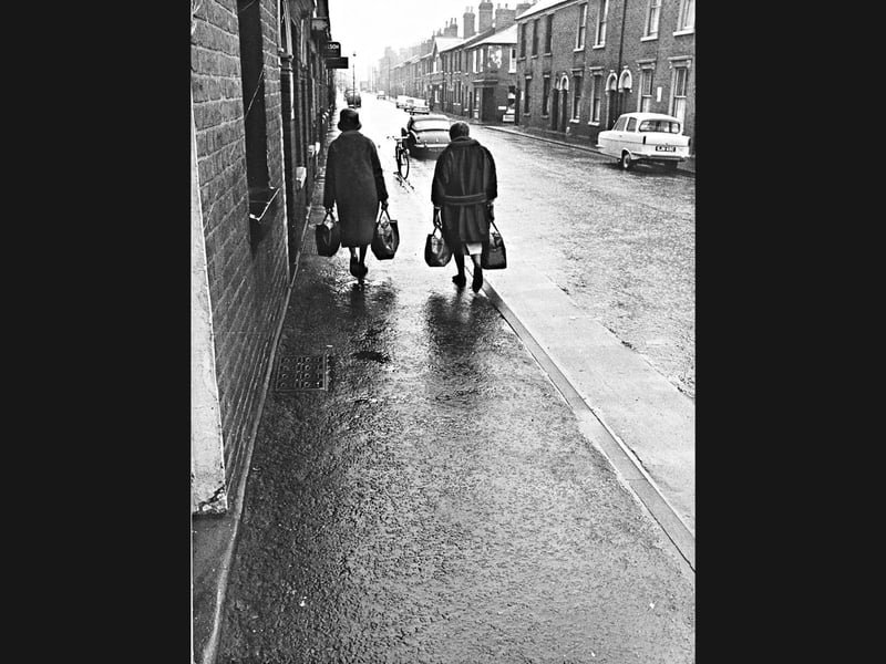 Two shoppers carry heavy bags along the rain-slicked pavement of Oak Street, Wolverhampton, in February 1966. Classic cars line the road alongside traditional terraced housing, capturing a quiet, everyday slice of mid-century life in the town. Do you remember Oak Street looking like this or recall doing your weekly shopping in the neighbourhood back in the 1960s?