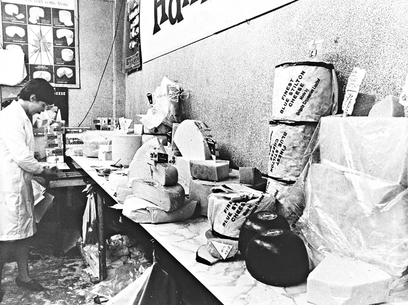 A worker tends to the vibrant counter display at Hawker's cheese stall in Wolverhampton in September 1966. The stall features an array of large wheels, blocks, and wrapped cheeses—including "Finest Blue Stilton made by Unigate"—alongside a educational wall poster displaying a map of regional English country cheeses and their origins. Do you remember visiting Hawker's stall or buying fresh regional cheeses in Wolverhampton back in September 1966