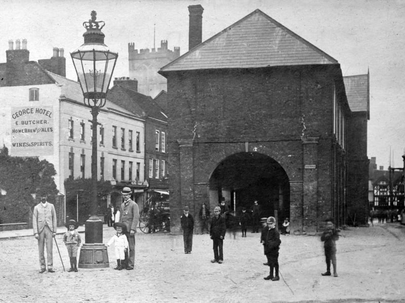 The old Ludlow Market Hall, aka Ludlow Town Hall, which was demolished in 1887. It was replaced with a new Ludlow Town Hall,  invariably said to have been built in 1887, and indeed the town hall carried a legend on the building declaring “Erected 1887”. It was strange then that when it was demolished in 1986 a time capsule was found under the foundation stone with a copy of the Ludlow Advertiser inside dated September 15, 1888. A conjectural explanation is that, as the town hall was intended to commemorate the golden jubilee of Queen Victoria of 1887, that date was trumpeted on the building, even though it appears not to have been built until the following year. Written in pencil on the back of this print is: "Old Town Hall. I think Mr Tyrrell (Town Clerk) in the white bowler.” 
