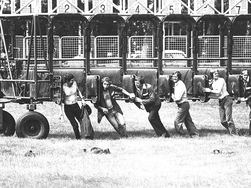 Straining muscles, grassy turf, and numbered metal gates, this July 1982 photograph captures stall handlers using sheer shoulder power to move the heavy starting stalls into place at Dunstall Park racecourse. With a race meeting about to get under way, the crew jumped inside the traps to manually guide the massive wheeled structure across the course. Some worked in open collared jackets while others went bare chested in the summer heat, offering an energetic, behind the scenes glimpse of the hard physical graft keeping Wolverhampton horse racing running smoothly. Do you remember spending a summer afternoon watching the horses run at Dunstall Park back in 1982? Who recalls seeing the grounds crew out on the course manhandling those heavy starting stalls into position?