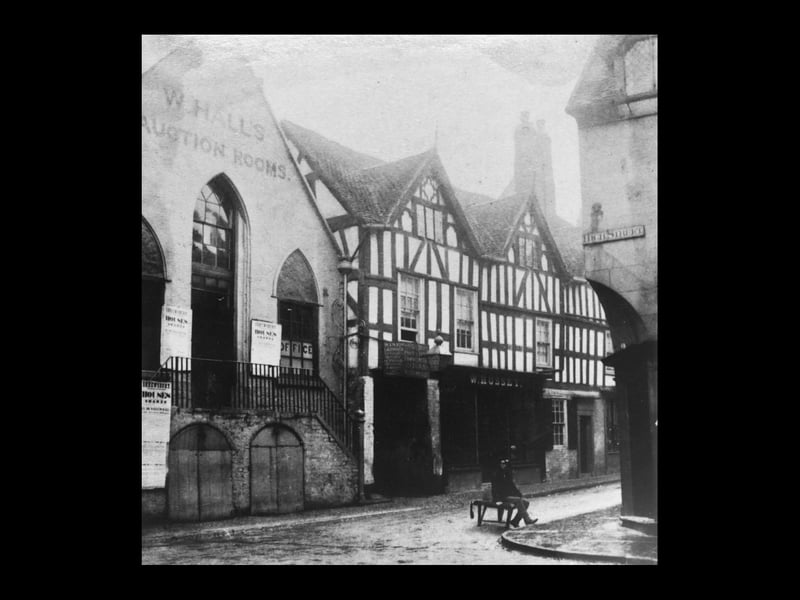 A man sits at the junction of High Street and Milk Street in Shrewsbury to have his shoes shined, and by staying still he has registered on the image while the shoe shiner, who was no doubt moving about, has not. The photo is thought to date from around 1866. Behind the man is W Hall's auction rooms, and "The Old Post Office Commercial Inn," with W Hussey next along.