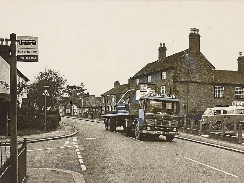 A heavy lorry rumbles along Main Street past picturesque timber-framed cottages and a Midland Red bus stop in Alrewas, near Lichfield, in January 1976. Local residents and Alrewas Conservation Society founder Councillor Margaret Stanhope voiced deep alarm that constant rumbling from heavy goods vehicles—including brewery lorries using the village as an alternative route—was cracking structural walls, loosening window frames, and damaging the foundations of historic buildings. Did you live along Main Street in Alrewas or remember the campaign to curb heavy freight traffic through the village during the winter of 1976?