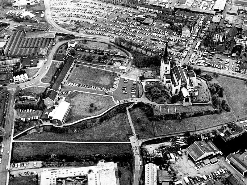 Captured from 1,500 feet during a helicopter flight organised for Walsall's planning committee, this sweeping aerial view shows the landmark spire of St Matthew’s parish church dominating the town's skyline in May 1974. The shot details the landscaped church lawns and gardens, the buildings of the Blue Coat school, packed car parks, and the bustling stalls of the open-air market stretching along High Street towards the top right. Can you pick out any familiar haunts or street corners from this high-altitude look over Walsall from May 1974?