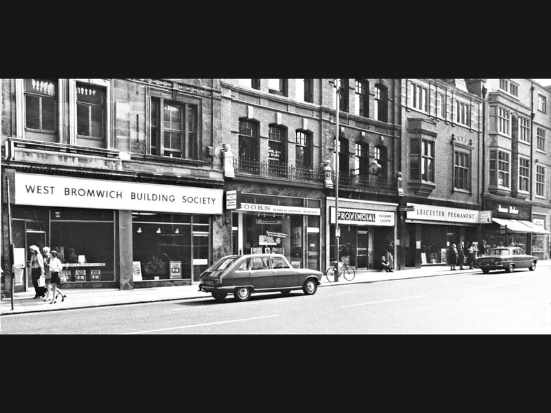 Shoppers stroll along the wide pavements of Lichfield Street in Wolverhampton on a bright summer afternoon in July 1974. A classic saloon car rests at the kerbside in front of a bustling row of local financial and commercial branches, featuring the prominent shopfront of the West Bromwich Building Society alongside Cooks World Travel Service, the Provincial Building Society, Leicester Permanent, and footwear retailer James Baker. Lined with ornate Victorian upper facades and neat mid-seventies signage, the thoroughfare served as one of the town centre's major commercial arteries. Do you remember calling into these shops on Lichfield Street or popping into town to manage your savings in Wolverhampton during the 1970s?