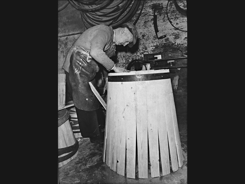 Surrounded by hoops and seasoned timber at Thomas Trevis Smith Ltd, cooper Joseph Gill expertly shapes a wooden barrel in March 1974. Captured in the heart of his workshop, Gill prepares to "fire" the barrel after hooping and setting one end, using heat to bend the staves so the loose flared ends can be drawn tightly together into their finished shape. The Cradley Heath firm kept alive a revered, centuries-old trade that supplied traditional wooden casks to breweries, chemical works, and local industries long after mass production had taken hold. Do you remember Thomas Trevis Smith Ltd or recall watching the skilled coopers at work in Cradley Heath back in 1974?